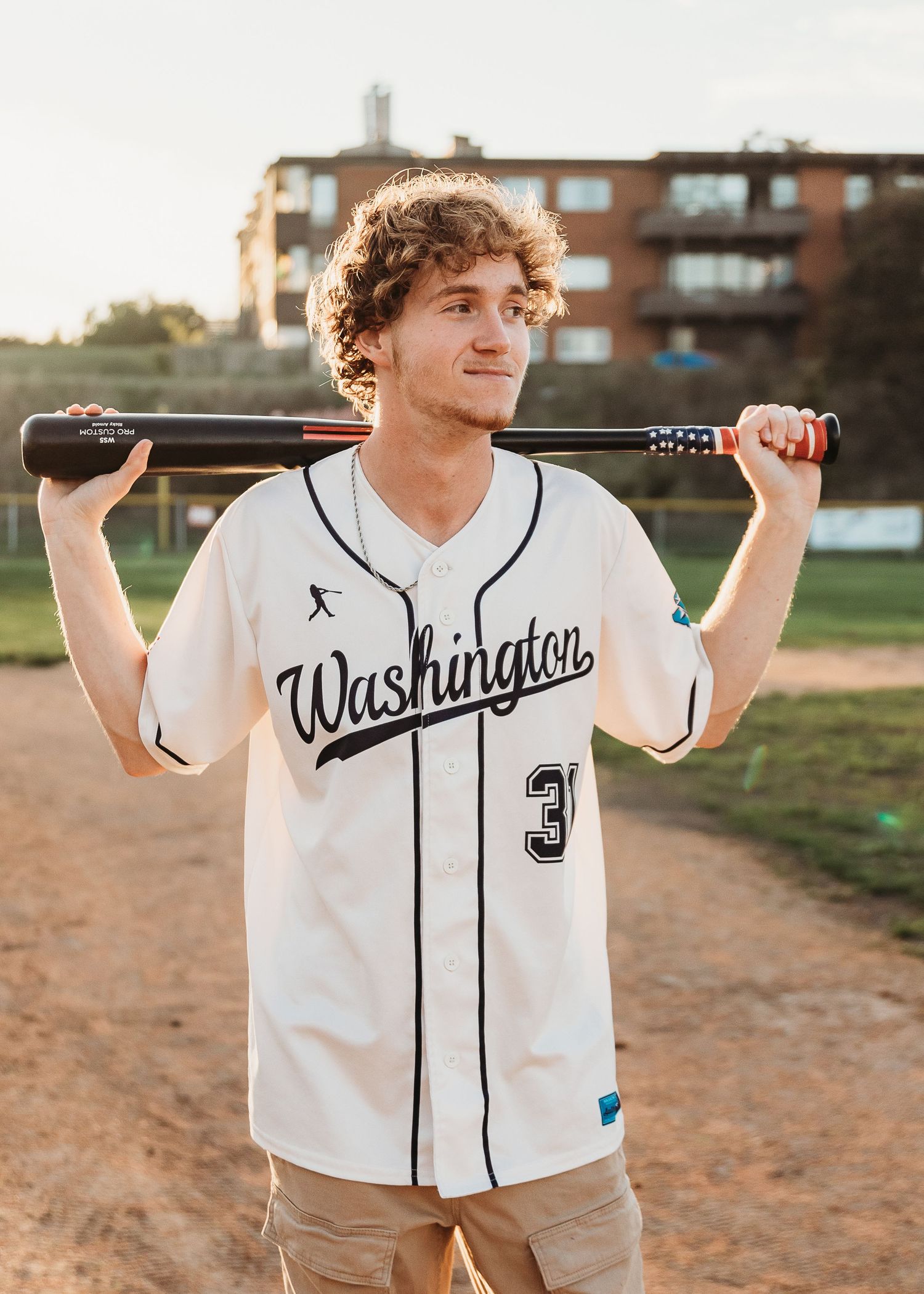 Teen baseball player in jersey holding glove stands before garage door in golden hour light.
