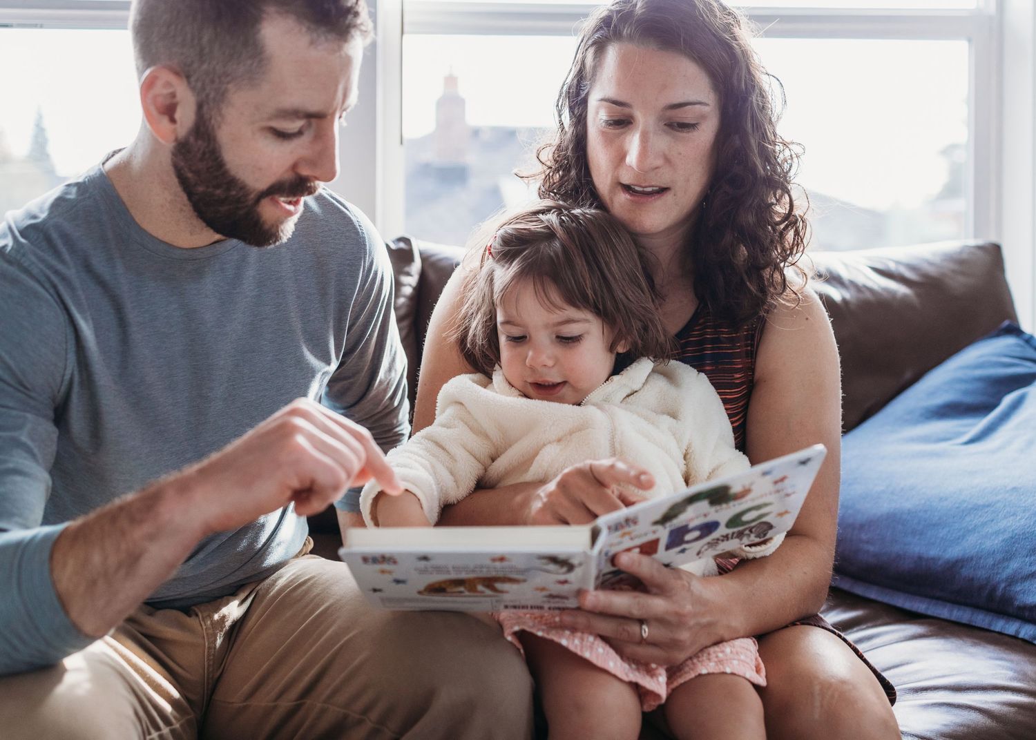 Parents and toddler reading a colorful picture book together on a cozy sofa at home.