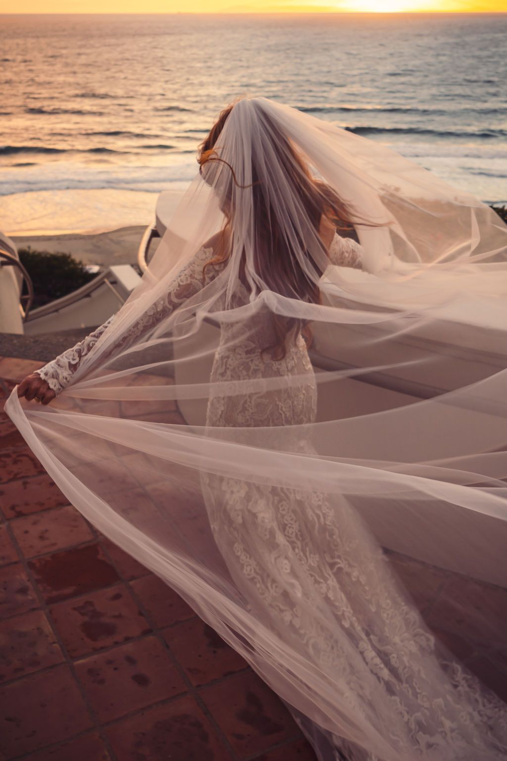 Bride in lace gown with flowing veil caught by ocean breeze at sunset on a coastal terrace.