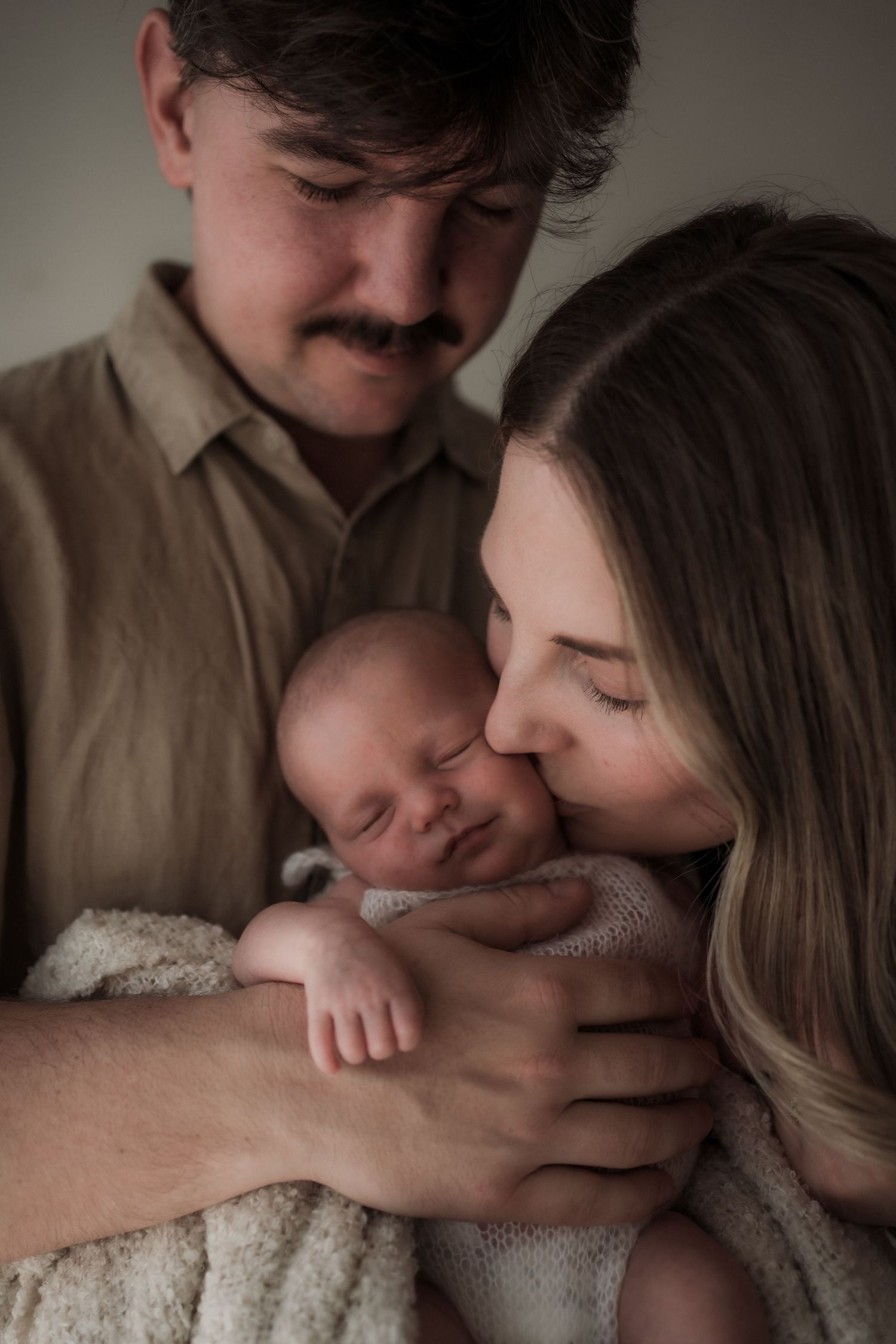 Parents tenderly hold and kiss their sleeping newborn baby wrapped in a soft white blanket in a studio portrait in the Yarra Valley