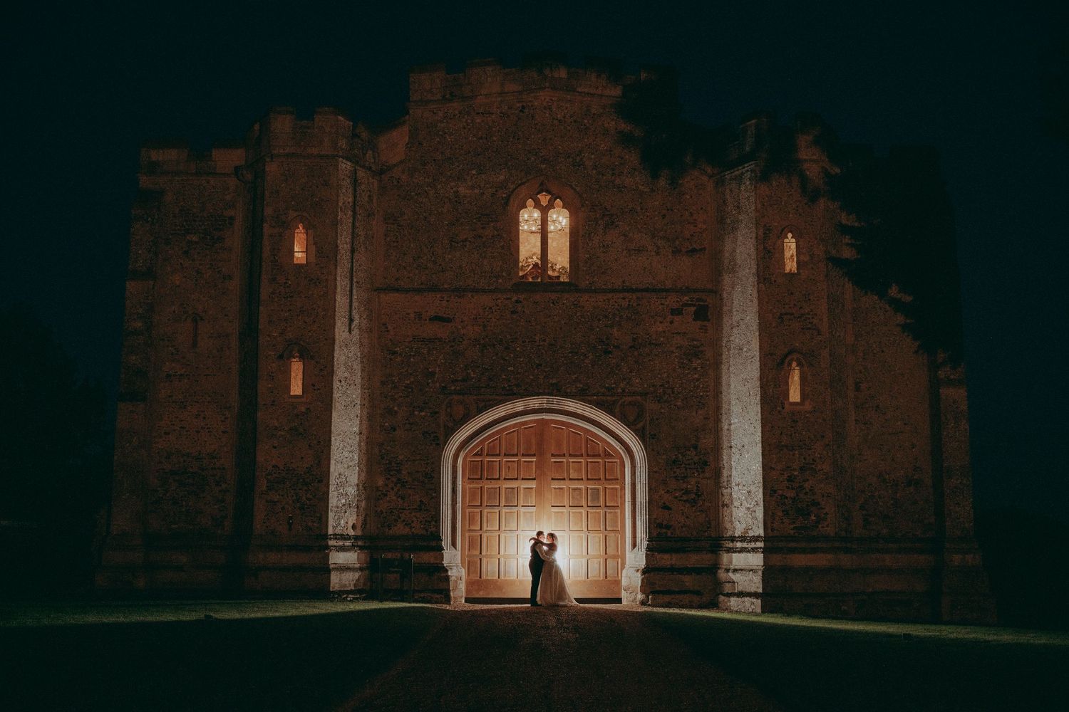 Couple silhouetted in glowing archway of historic brick castle at night during romantic wedding ceremony.
