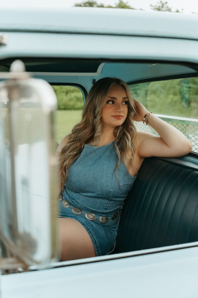 Young woman in denim outfit sitting in vintage car backseat, posing with long wavy hair and a confident smile.