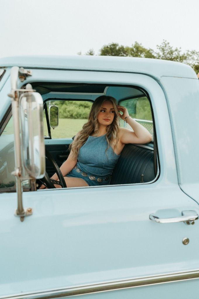 Young woman in denim outfit sitting in vintage light blue pickup truck cab, posing for senior photos.