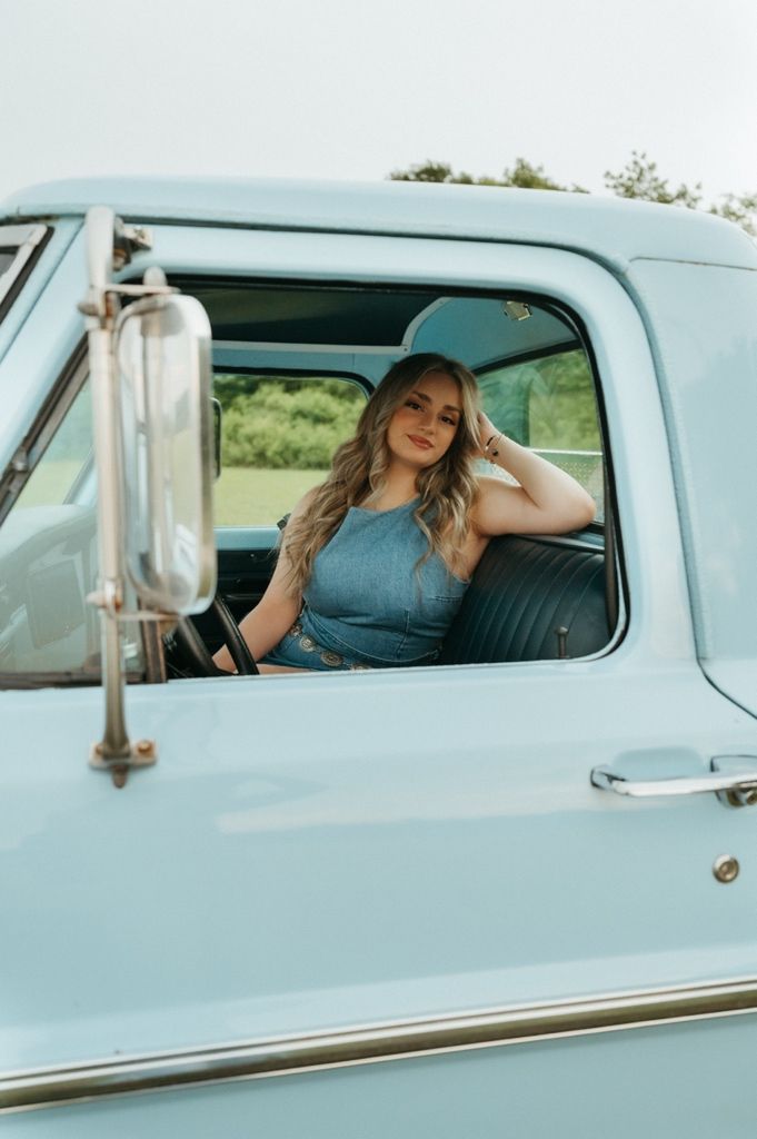 Young blonde woman in blue dress sitting in vintage light blue pickup truck with green trees in background.