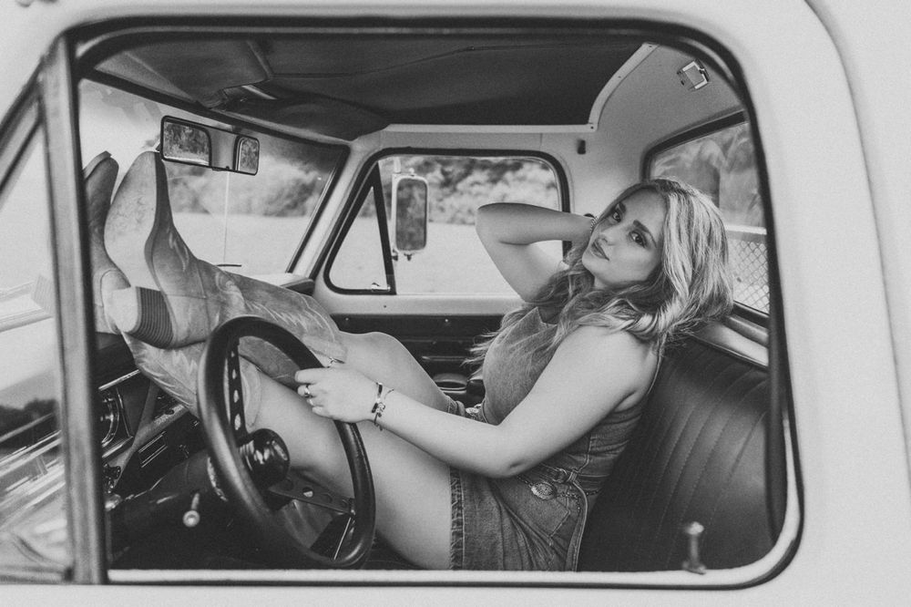 Black and white photo of a blonde woman posing in a vintage truck cab, wearing boots and a denim outfit.