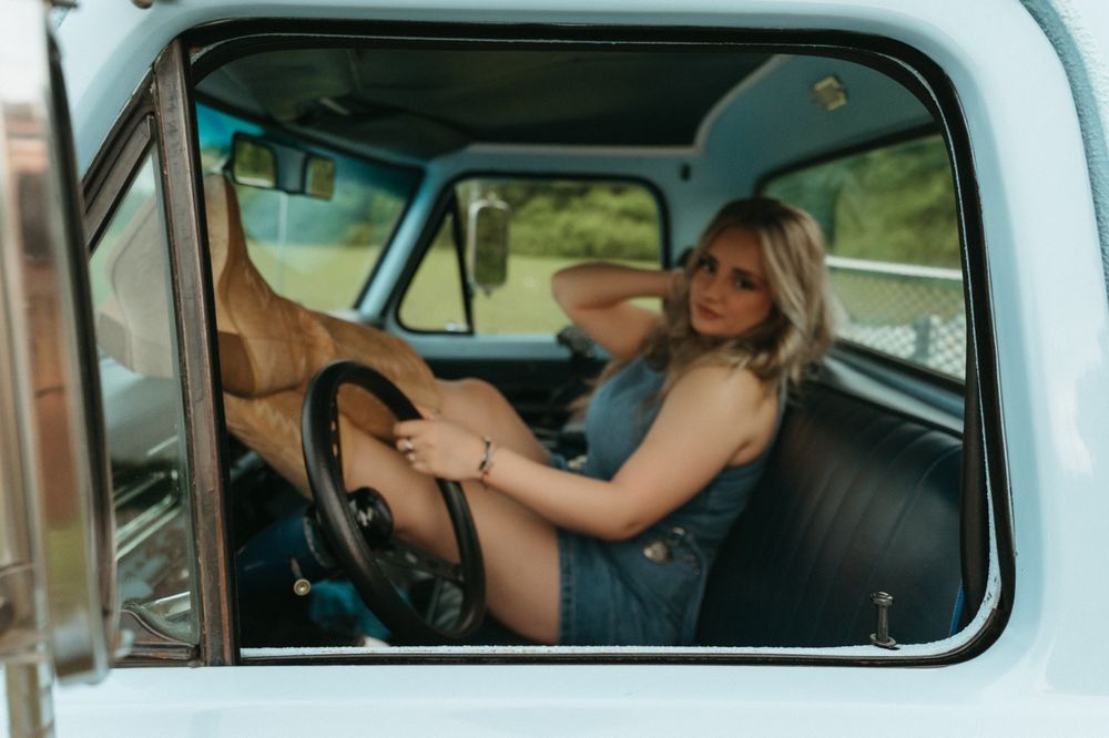 Woman in denim outfit resting cowboy boots on steering wheel of vintage light blue truck.