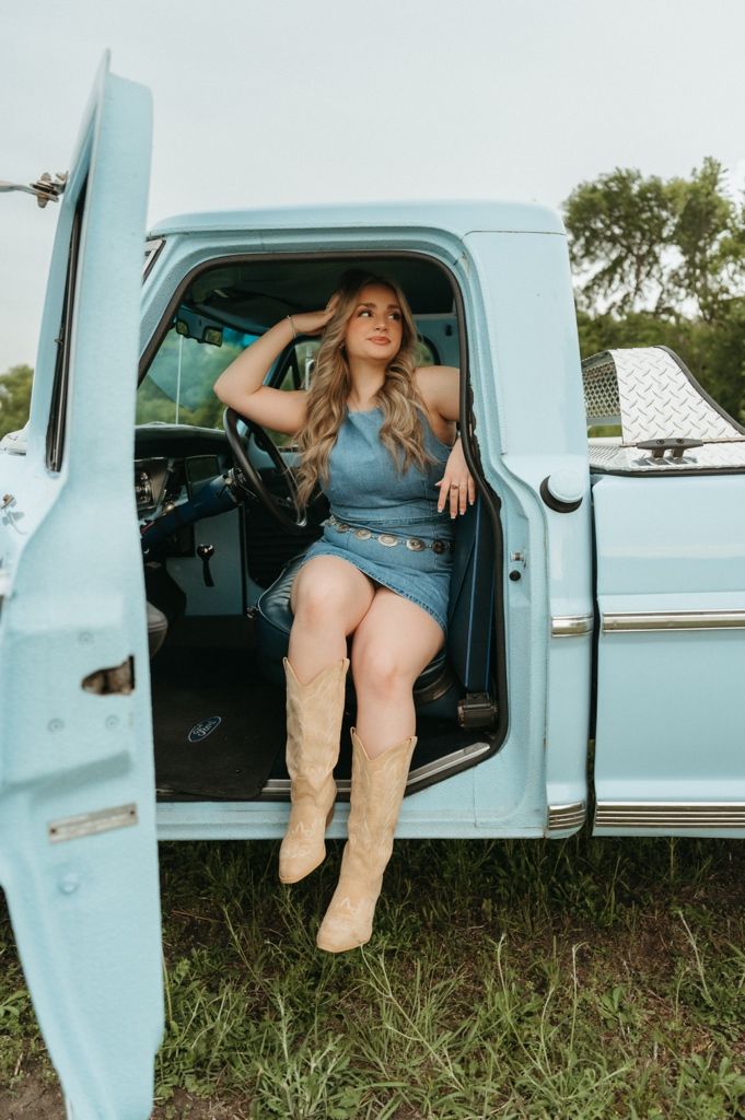 Woman in denim dress and tan cowboy boots sitting in open door of vintage light blue pickup truck outdoors.