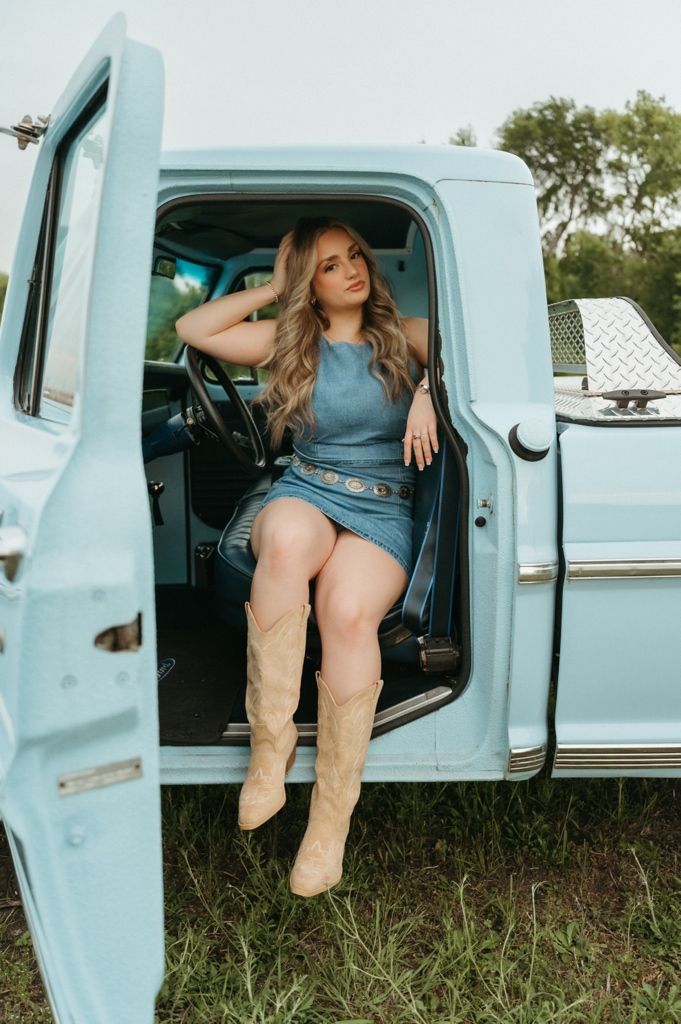 Young woman in denim dress and cowboy boots posing in vintage light blue pickup truck door.