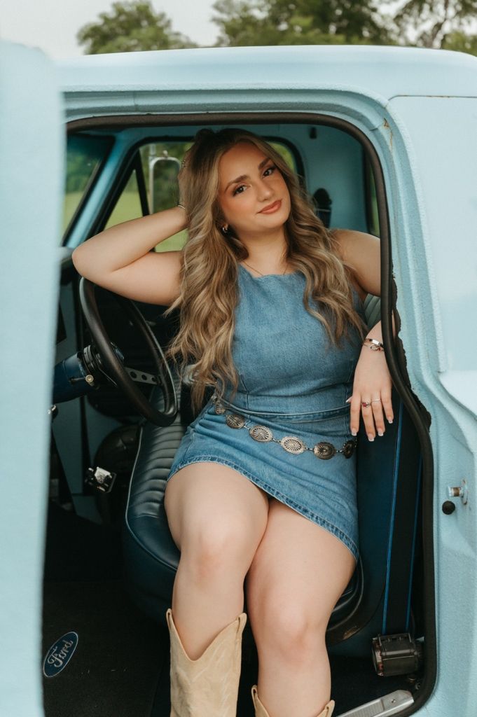 Woman in denim dress and cowboy boots posing in a vintage light blue Ford truck for a country-style photo shoot.