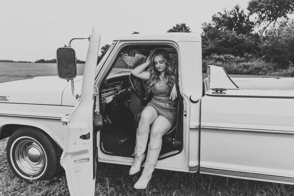 Black and white photo of a blonde woman in boots sitting in the open door of a vintage pickup truck in a field.