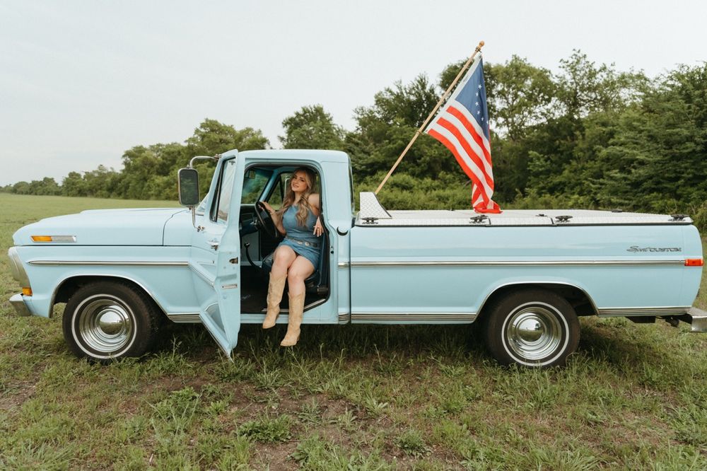 Woman sitting in a vintage light blue pickup truck with an American flag in a green field.