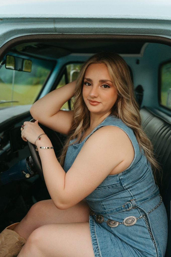 Young woman in denim dress with silver belt posing in vintage truck cab.