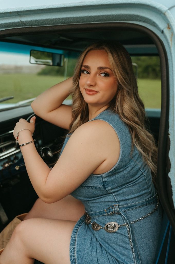 Blonde woman in denim outfit smiling over shoulder in classic truck interior.