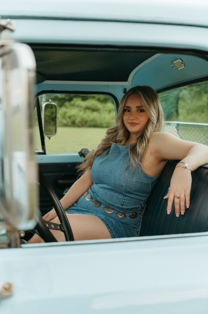 Smiling blonde woman in denim romper sitting inside classic light blue vintage pickup truck.
