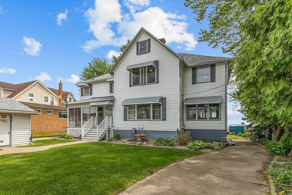 White two-story colonial home with blue accents, covered porch, and lush green lawn under a bright blue sky.