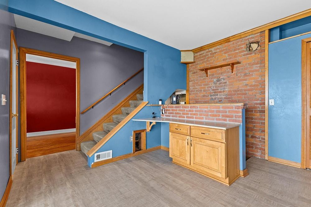 Kitchen area with blue walls, brick accent wall, wooden cabinets, and staircase with red door.