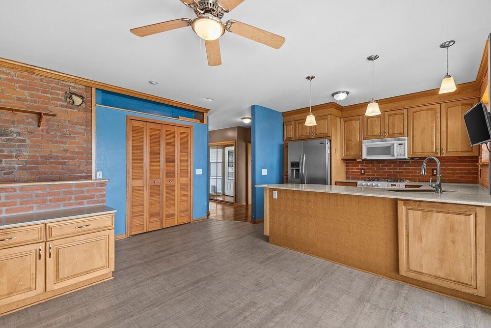Open kitchen with oak cabinets, stainless steel appliances, brick accent wall, and hardwood floors under a ceiling fan.