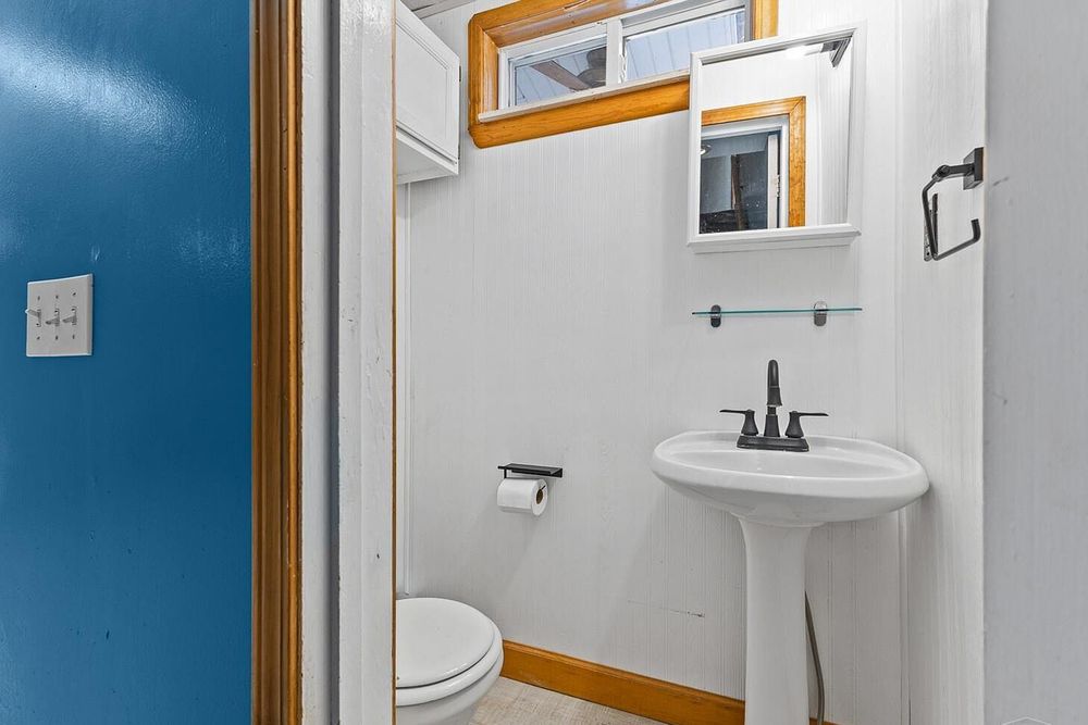 Small half bathroom with white pedestal sink, toilet, wood trim accents, and a window above the mirror shelf.