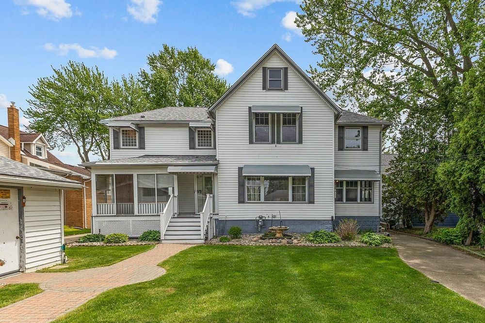 White two-story colonial home with covered porch, dormers, and lush green lawn surrounded by mature trees.