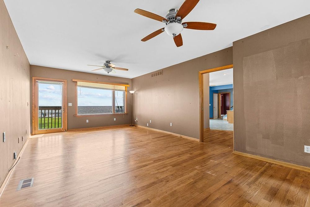Bright empty living room with hardwood floors, ceiling fan, neutral tan walls, and large windows offering outdoor views.