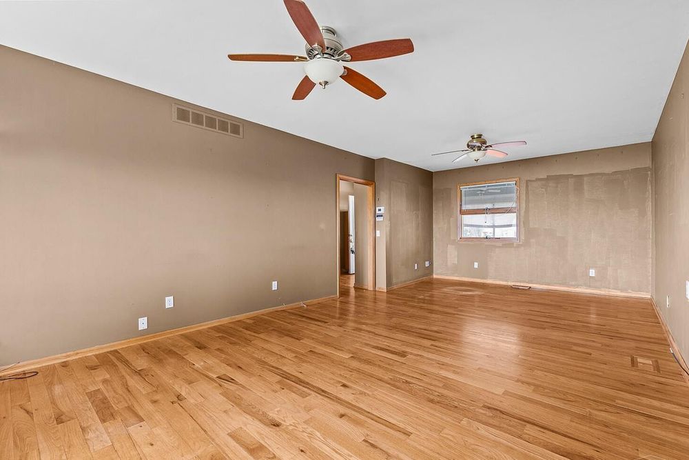 Spacious empty living room with light hardwood floors, neutral beige walls, and a ceiling fan with light fixture.