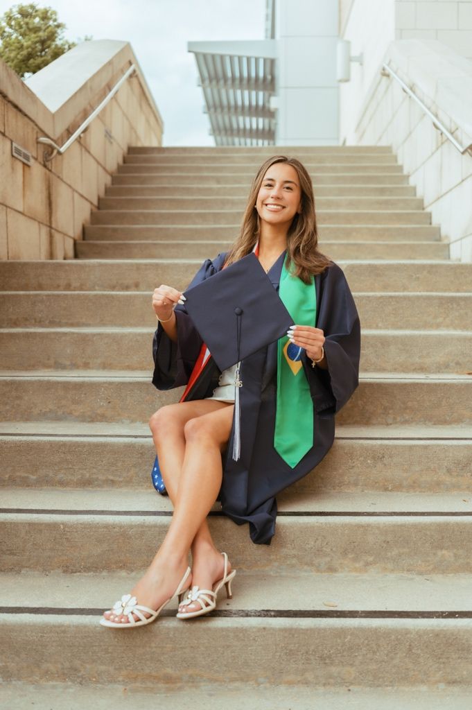 Happy graduate in navy cap and gown with green Brazilian flag stole sitting on campus steps.