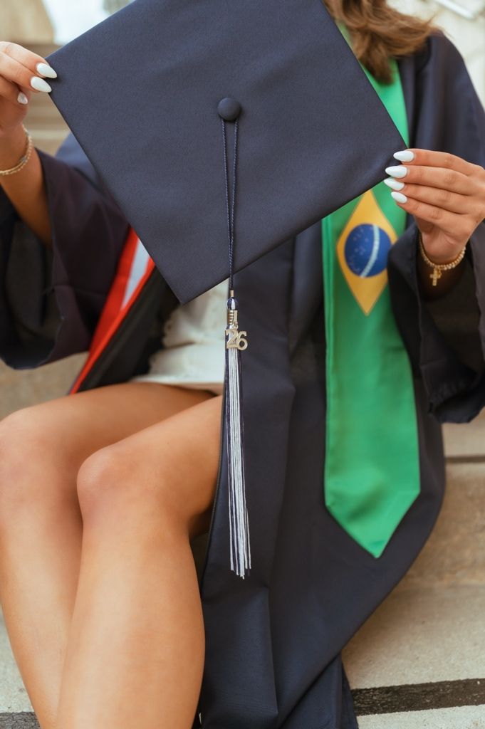 Close-up of graduate holding navy mortarboard cap with tassel wearing Brazilian flag graduation stole.