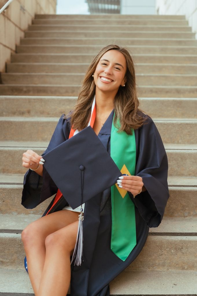 Smiling female graduate in navy cap and gown with green sash sitting on outdoor steps holding graduation cap.