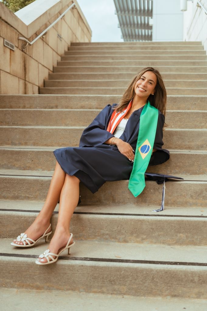 Smiling graduate in navy gown with Brazilian flag stole sitting relaxed on outdoor stone steps.
