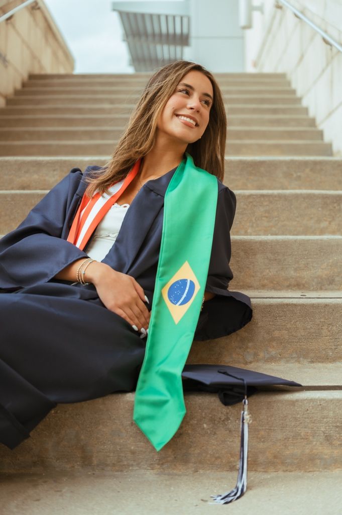 Female graduate sitting on stairs wearing black gown with Brazilian flag honor stole.