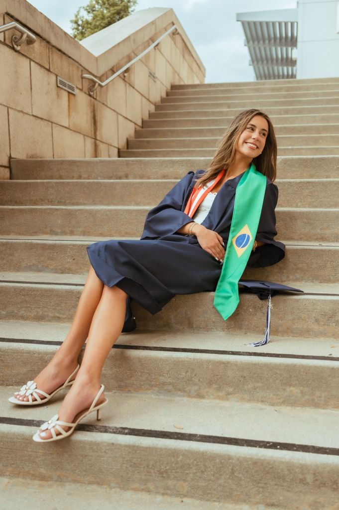 Female graduate in navy gown with Brazilian flag sash sitting relaxed on outdoor concrete steps.