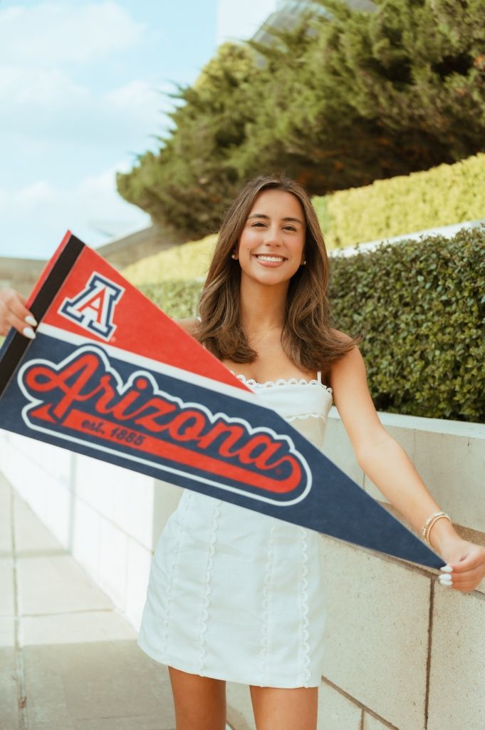 Smiling young woman holding an Arizona Wildcats pennant flag outdoors, wearing a white dress.
