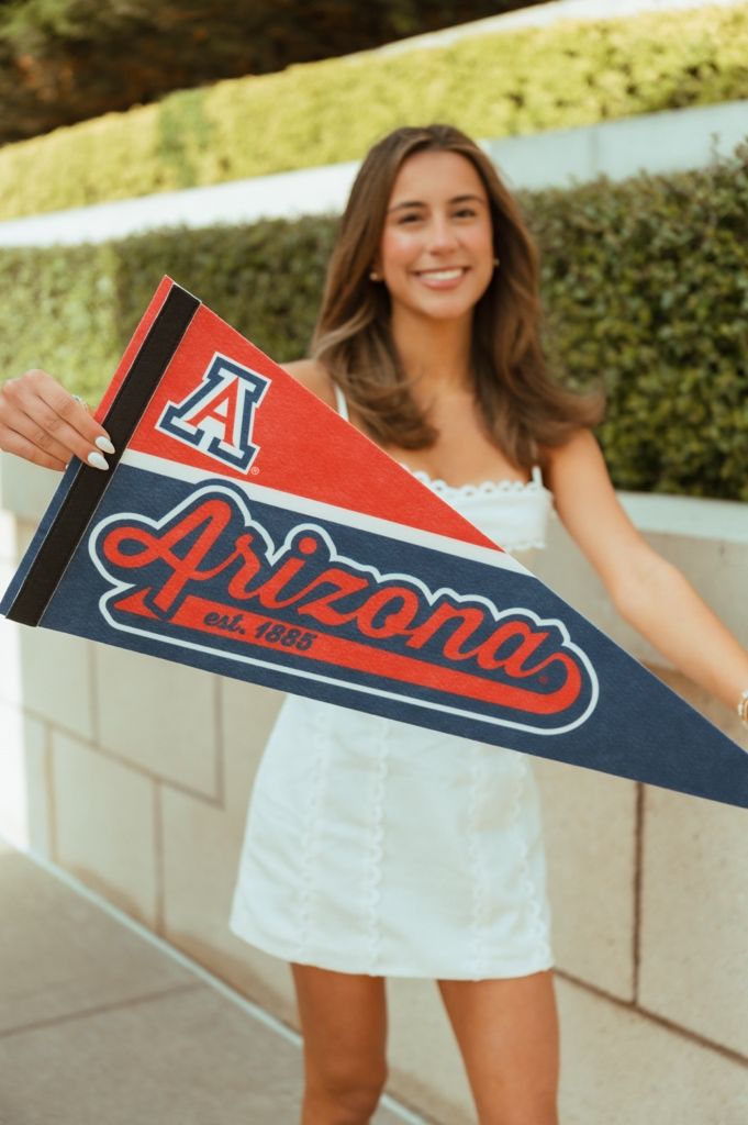 Smiling young woman holding a University of Arizona pennant flag, wearing a white dress outdoors.