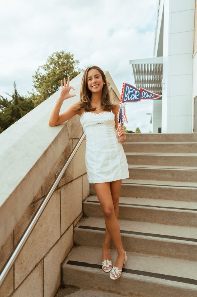 Smiling young woman in white mini dress holding pennant flags on outdoor stairs.