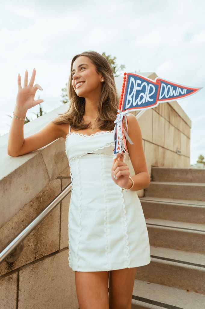 Smiling young woman in white mini dress holding a Bear Down pennant flag on outdoor steps.
