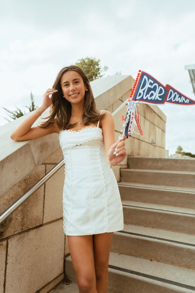 Young woman in white dress holding a Bear Down pennant flag on outdoor stairs at University of Arizona.