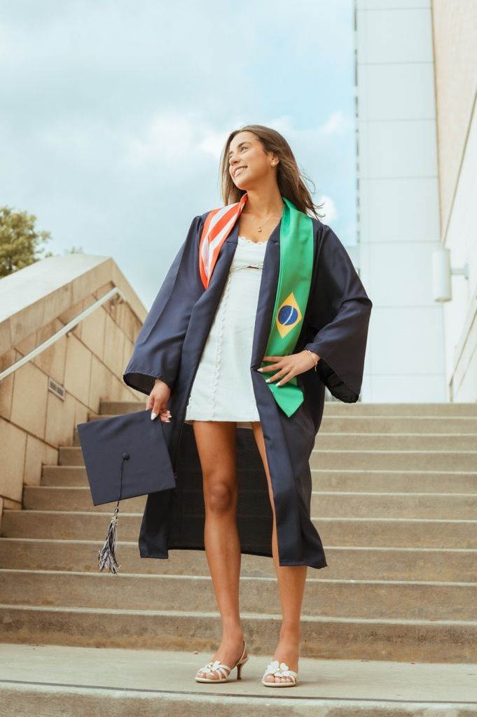Young woman in navy graduation gown with Brazilian flag stole holding mortarboard on outdoor steps.
