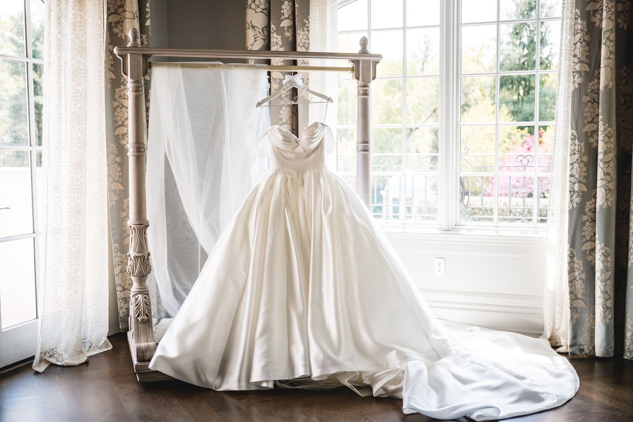 Elegant white ball gown wedding dress displayed on a wooden hanger in a bright, sunlit room with floral curtains.