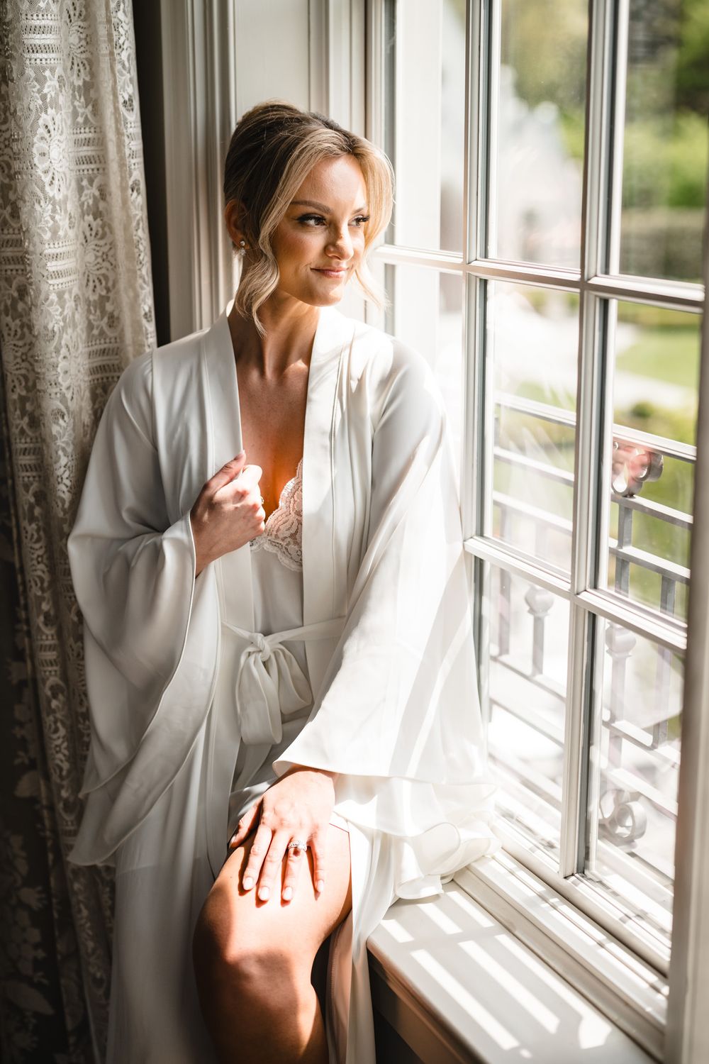 Bride in white silk robe sitting by sunlit window, elegant updo hairstyle, soft natural light.