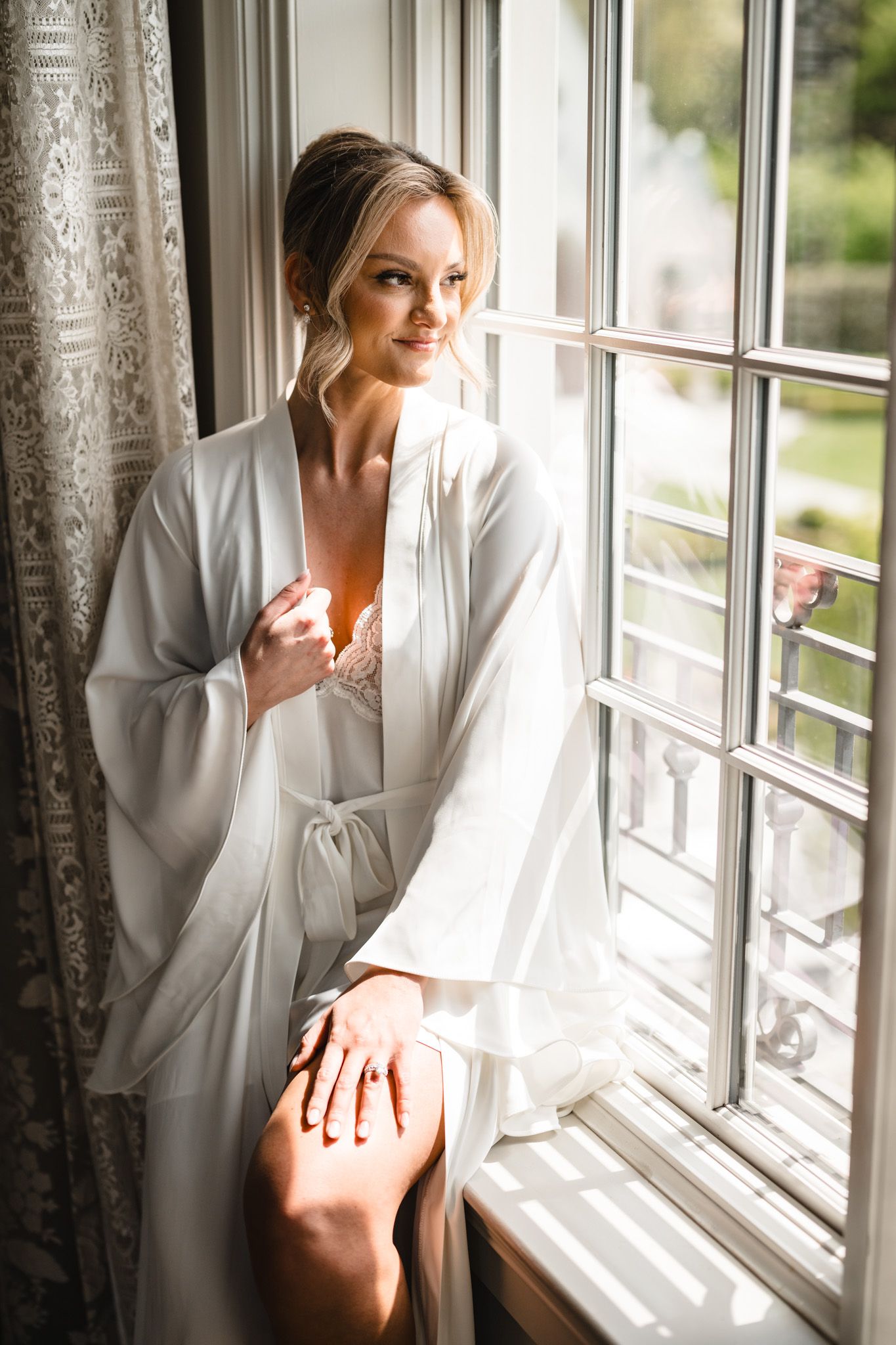 Bride in white silk robe sitting by sunlit window, elegant updo hairstyle, soft natural light.