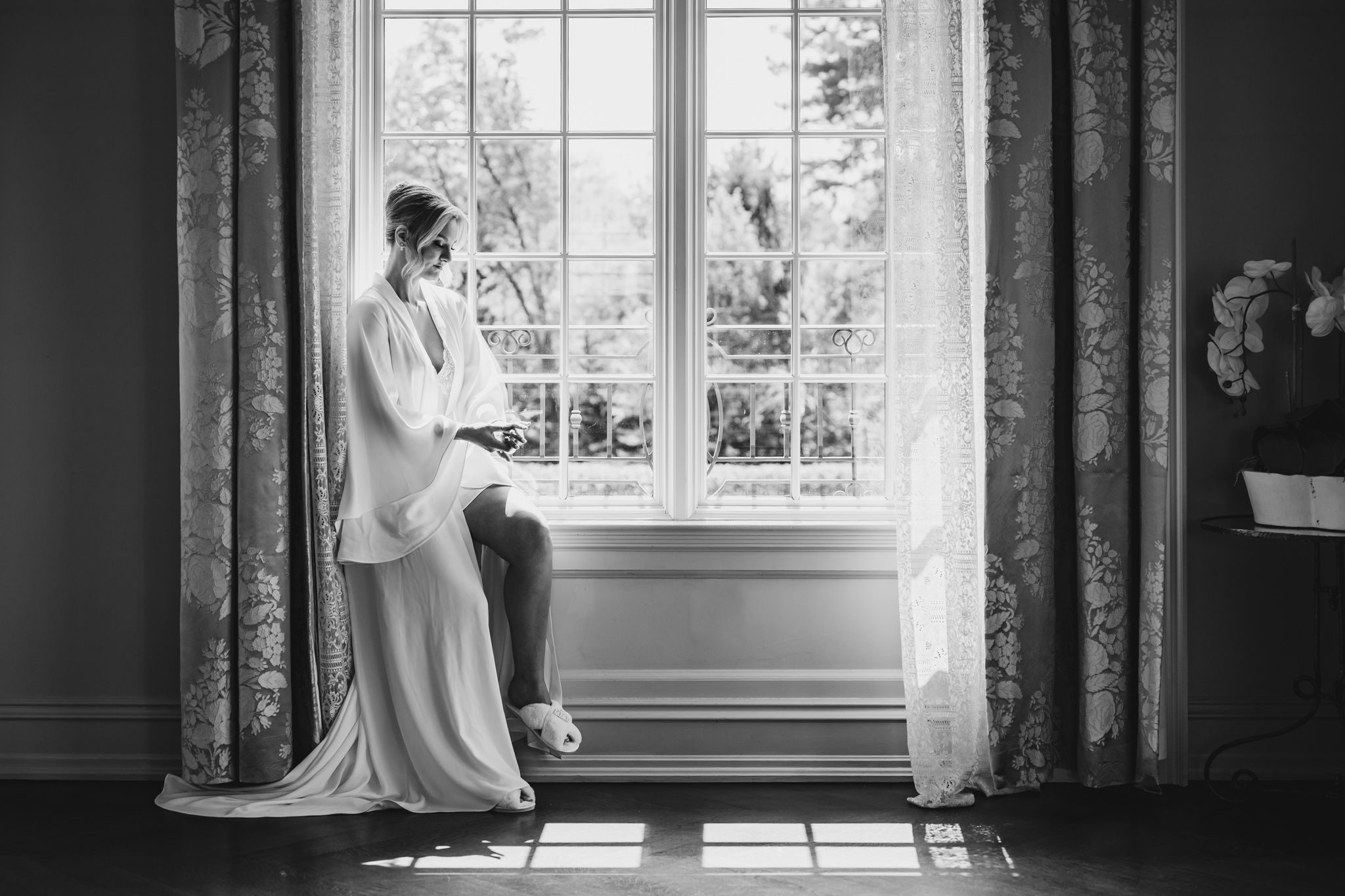 Black and white photo of a bride in a white gown sitting by a sunlit window with floral curtains.