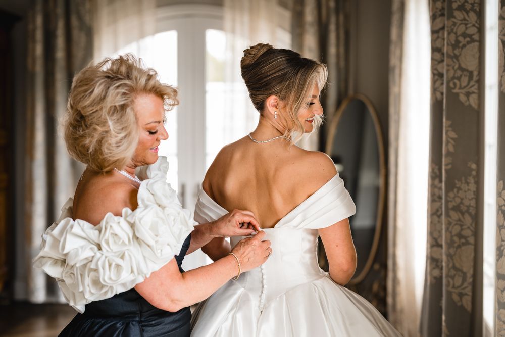 A woman helps a bride fasten her white off-shoulder wedding gown in an elegant room with a mirror.