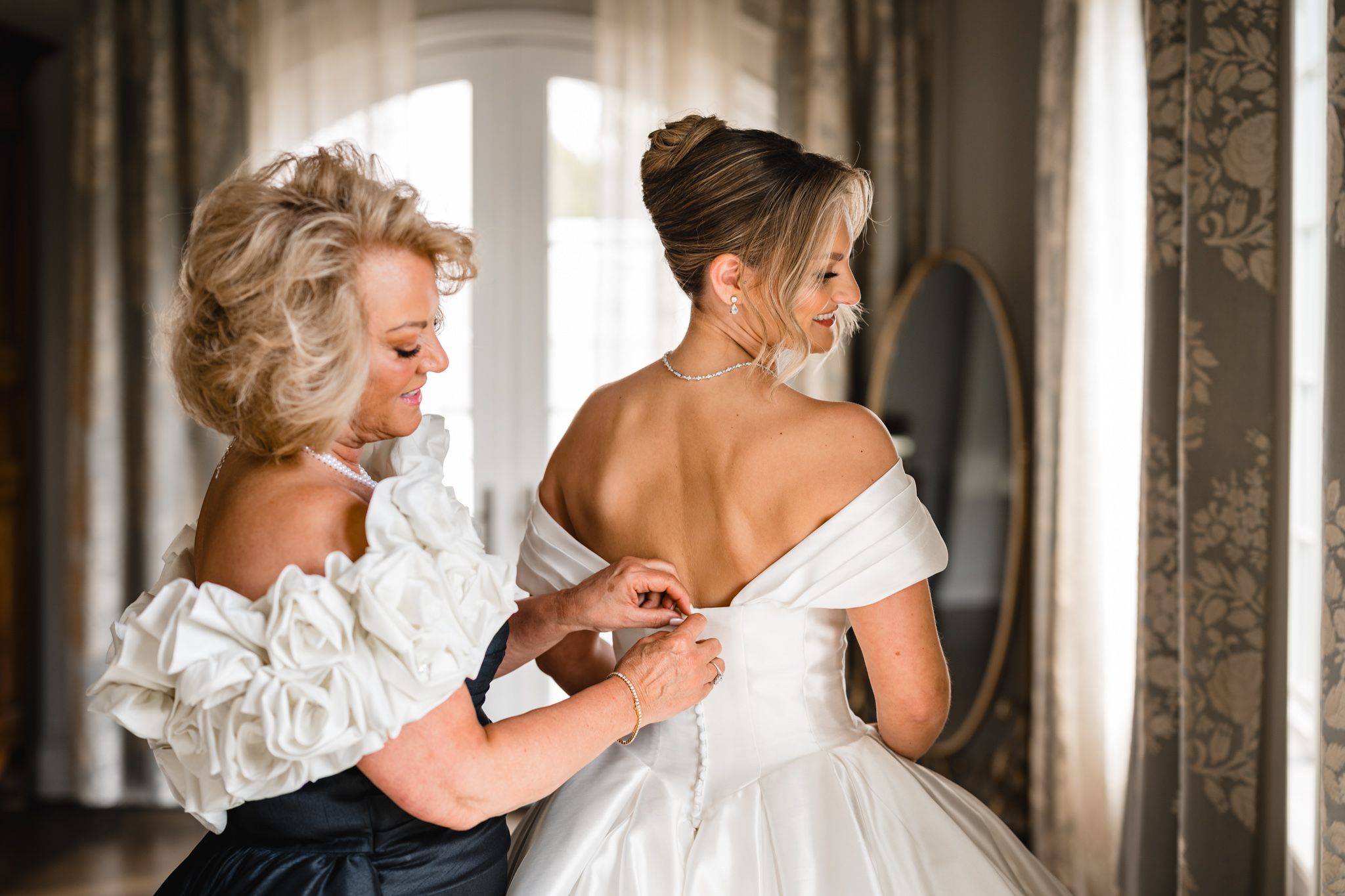 A woman helps a bride fasten her white off-shoulder wedding gown in an elegant room with a mirror.
