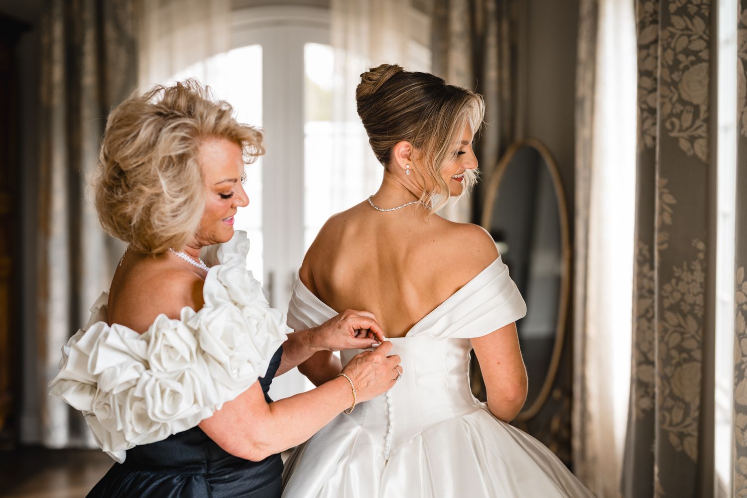 A woman helps a bride fasten her white off-shoulder wedding gown in an elegant room with a mirror.