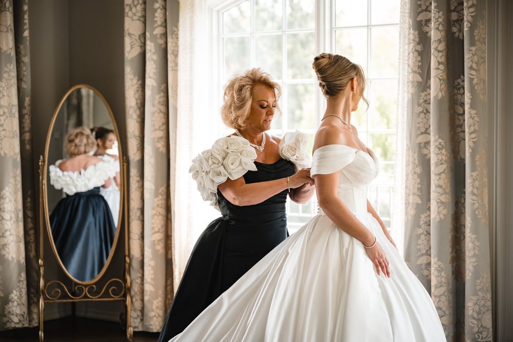 A mother helps her daughter into a white off-shoulder ball gown wedding dress near a window with a mirror reflection.