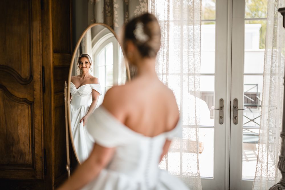 Bride in off-shoulder white gown admiring her reflection in an elegant oval mirror before her wedding.