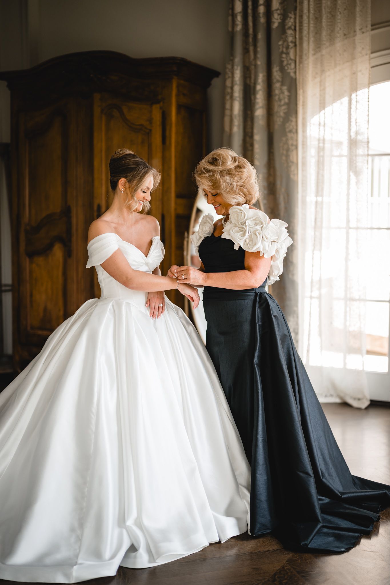 Bride in white ball gown getting help from woman in black dress near window.