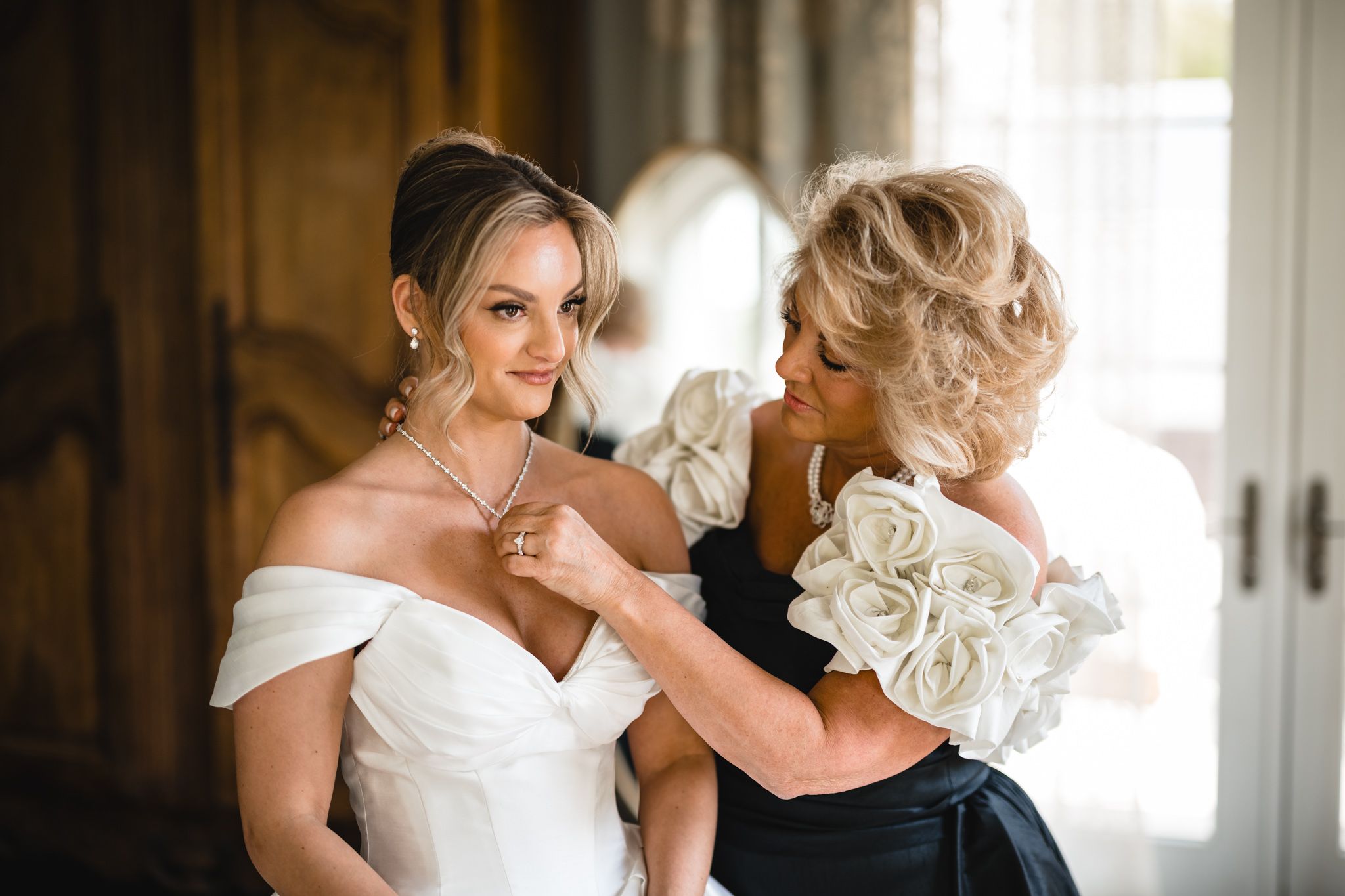 Bride smiling as older woman helps fasten necklace, both elegantly dressed in white and black gowns.