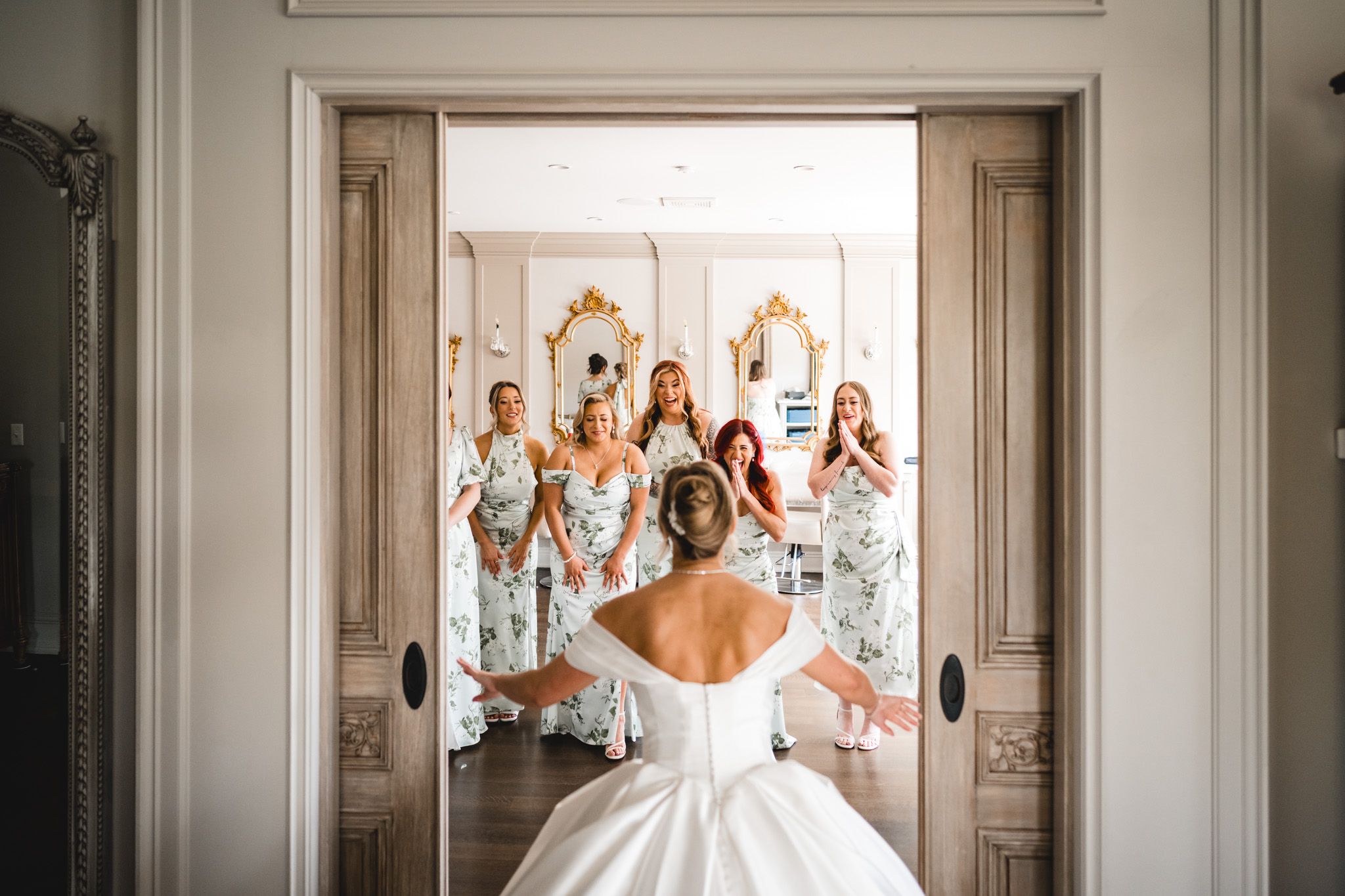 Bride in white ball gown reveals herself to excited bridesmaids in floral dresses through elegant doorway.
