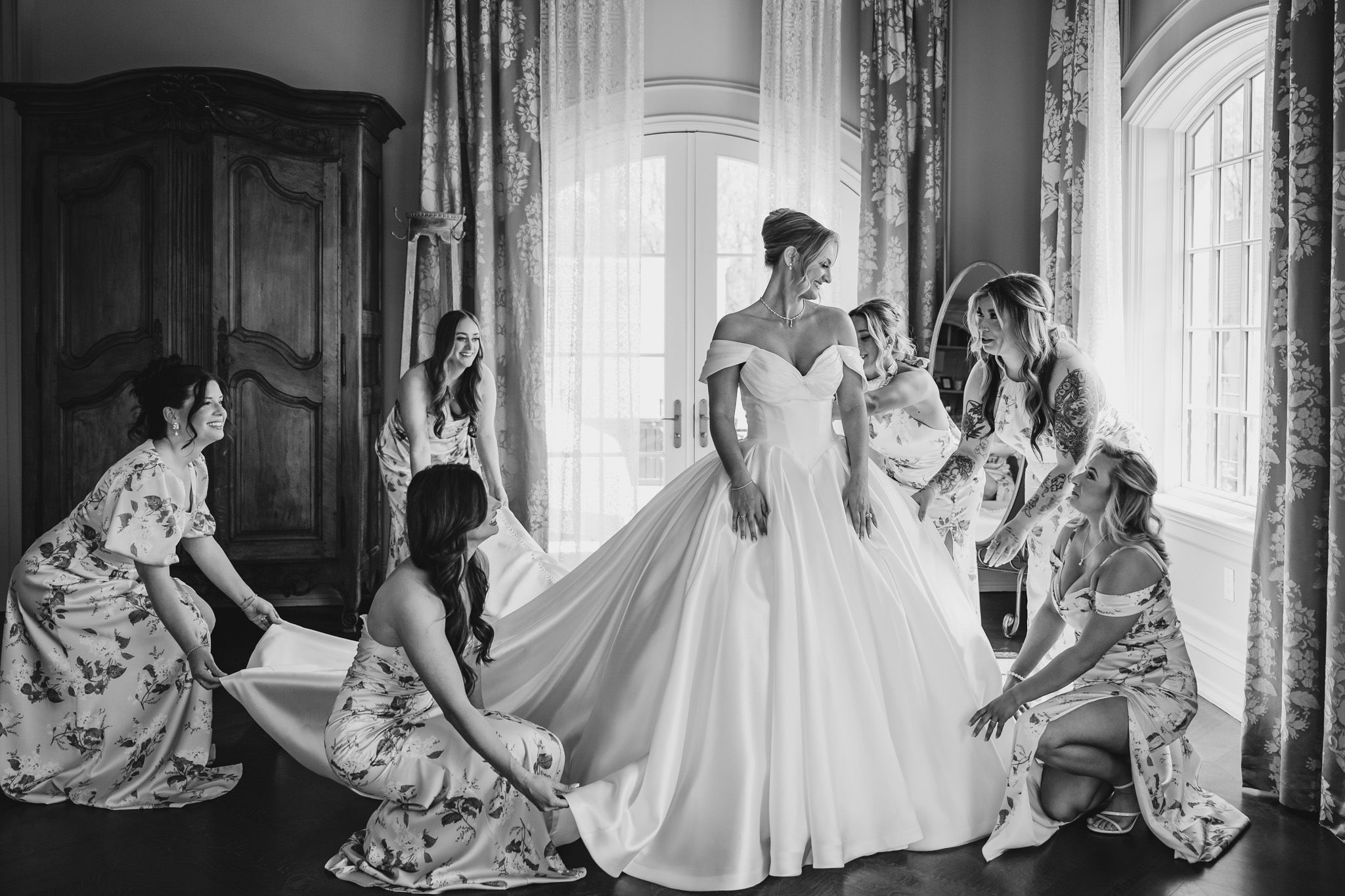 Bridesmaids in floral dresses helping bride with white ball gown in black and white photo.